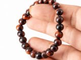 Fingers holding a genuine natural Red Tiger Eye gemstone bracelet vertically, showcasing the 8mm beads.