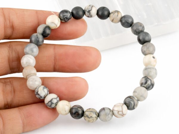 Fingers gently holding a genuine natural Spider Web Agate gemstone bracelet, showing the elastic cord and intricate grey and white 8mm beads.