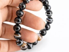 Fingers gently holding a genuine natural Sulemani Hakik gemstone bracelet vertically, showing the elastic cord and striking multi-colored banded 8mm beads.