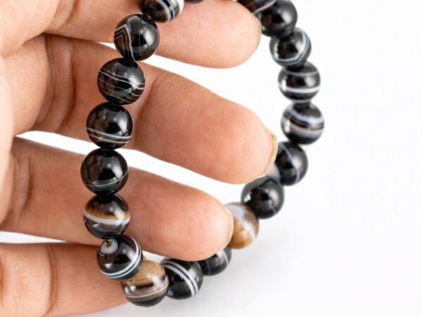 Fingers gently holding a genuine natural Sulemani Hakik gemstone bracelet vertically, showing the elastic cord and striking multi-colored banded 8mm beads.